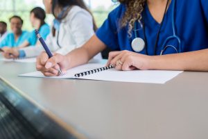 An unrecognizable medical student sits at a table in her lecture hall and writes in her class workbook.